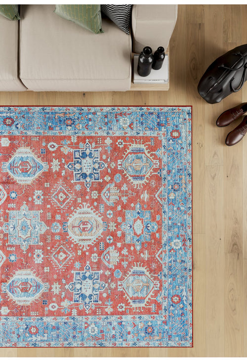 Load image 1 in gallery view. The Lunavale Red Blue Washable Rug - LCC3007 (Custom Size), featuring red, blue, and cream geometric patterns, sits on a light wooden floor beside a beige sofa—ideal for high-traffic areas. Shoes, a bag, and water bottles rest nearby.