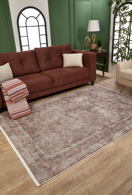 Load image 1 in gallery view. A living room with a brown sofa, beige pillows, a striped blanket, and the Luxe Borderline - Washable Rug - JR1741 (Custom Size) in bronze and brown—ideal for busy spaces—plus a small lamp, wooden floor, green paneled walls, and an arched window.