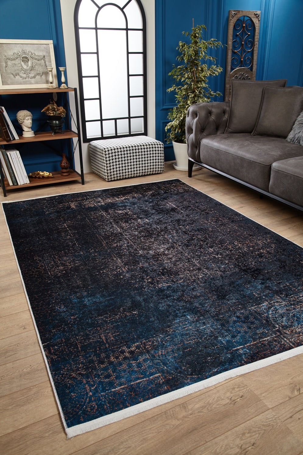 A cozy living room with the Luxurious Fleur - Washable Rug - JR1897 (Custom Size), perfect for high-traffic areas, paired with light wood flooring, a gray tufted sofa, potted plant, bookshelves, black and white ottoman, and an arched window.