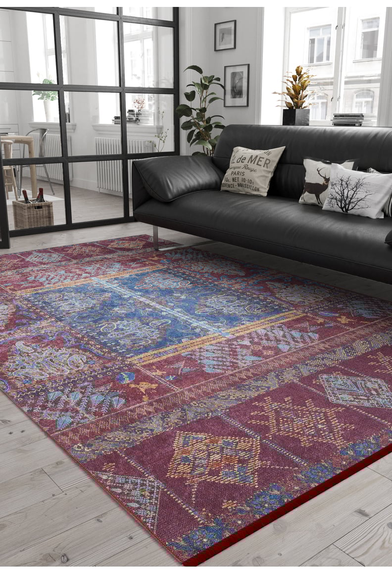 A modern living room with a black sofa, printed pillows, and lush plants features the Majestic Mandarin - Washable Rug - JR1863, ideal for high-traffic areas on a light wooden floor, with large windows letting in natural light.