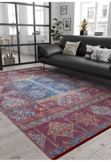 A modern living room with a black sofa, printed pillows, and lush plants features the Majestic Mandarin - Washable Rug - JR1863, ideal for high-traffic areas on a light wooden floor, with large windows letting in natural light.