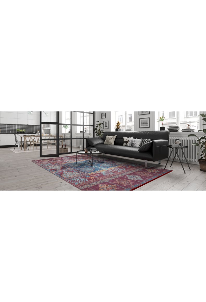 A modern living room features a black sofa, patterned cushions, a round coffee table, and the Majestic Mandarin - Washable Rug - JR1863 in colorful geometric patterns—ideal for busy spaces. Large windows provide natural light and a dining area is visible in the background.