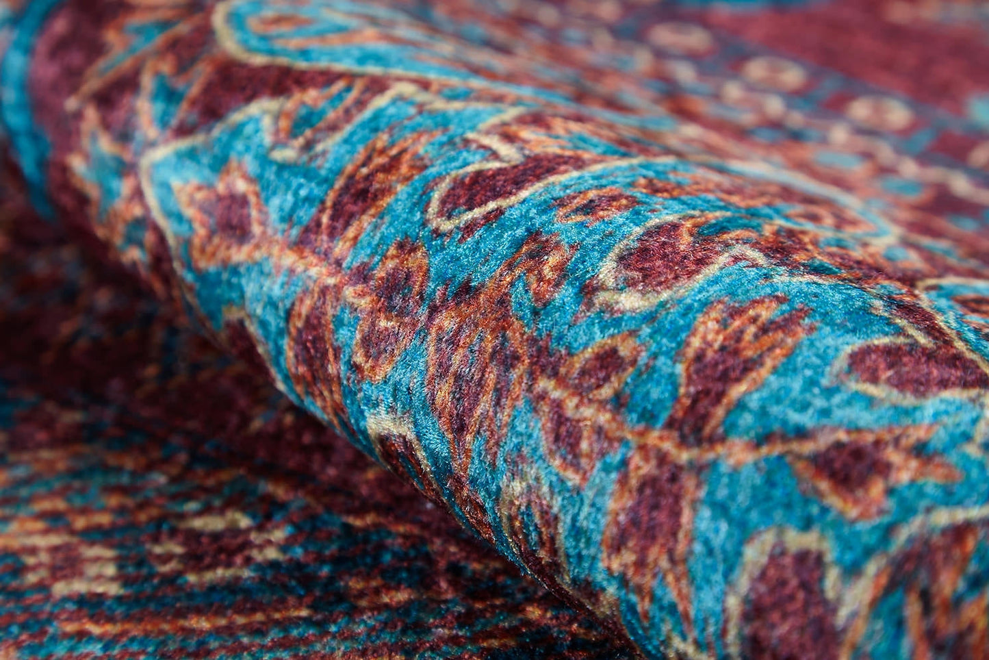 A close-up of the Majestic Mandarin - Washable Rug - JR1863 (Custom Size) shows its textured red weave with intricate blue and gold geometric patterns. Partially rolled, it highlights vibrant fibers—perfect for high-traffic spaces.