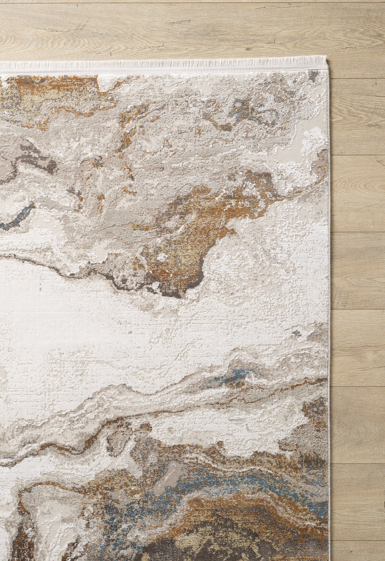 A corner of the Majestic Marble Premium Area Rug - M673M, featuring a marbled abstract design in cream, brown, gray, and blue with a thin fringe, rests on a light wood floor.