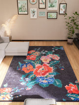 A modern living room with a light gray sectional, wood flooring, framed botanical prints, potted plants in the corners, and the Meadow Dream - Washable Rug - JR2001—a black rug with vibrant red, blue, and green floral patterns ideal for high-traffic areas.