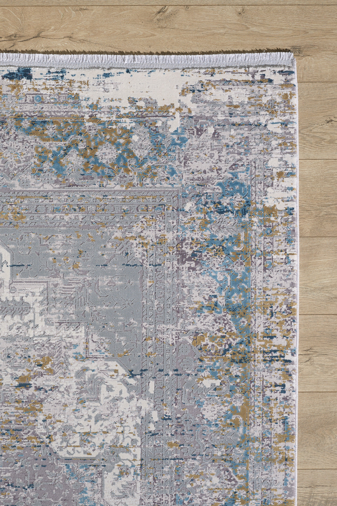 A section of the Medallion Revamp Modernised Medallion Rug - MT002, in blue, gold, gray, and white with a distressed abstract design and fringed edge, lies on a light wood floor.