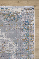 A section of the Medallion Revamp Modernised Medallion Rug - MT002, in blue, gold, gray, and white with a distressed abstract design and fringed edge, lies on a light wood floor.