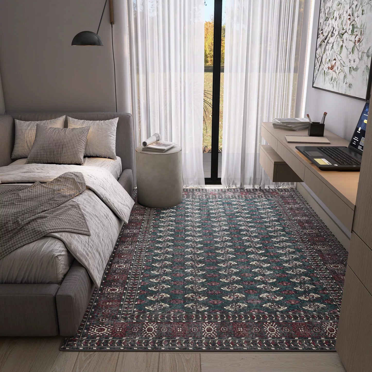 A modern bedroom with a gray bed, pillows, throws, a Midnight Tribal - Washable Rug - JRN1034 perfect for high-traffic areas, a round gray ottoman, desk with laptop, wall lamp, and large windows with sheer white curtains letting in light.