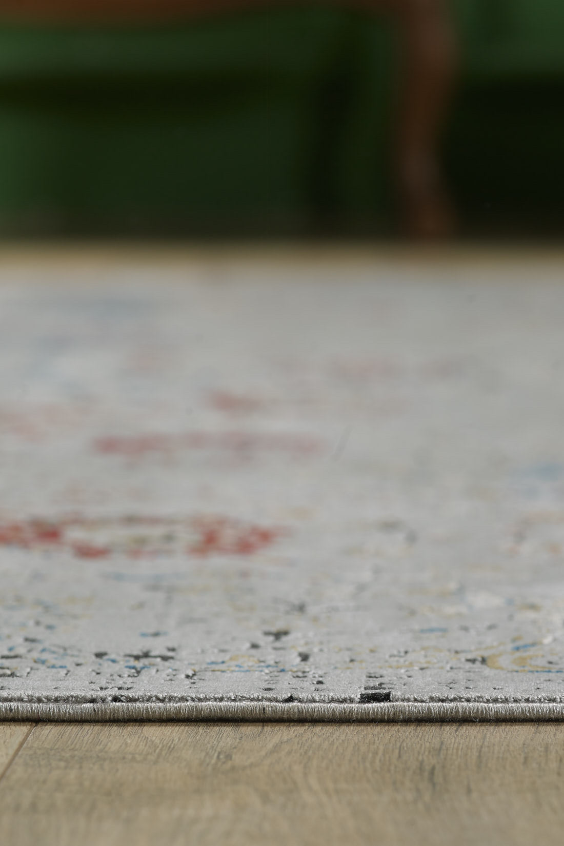 Close-up of the edge of the Miniature Elegance Oushak Rug - Grey - 2084F, a light-colored, machine-made rug with subtle patterns on a wooden floor; a blurred green object is visible in the background.