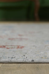 Close-up of the edge of the Miniature Elegance Oushak Rug - Grey - 2084F, a light-colored, machine-made rug with subtle patterns on a wooden floor; a blurred green object is visible in the background.
