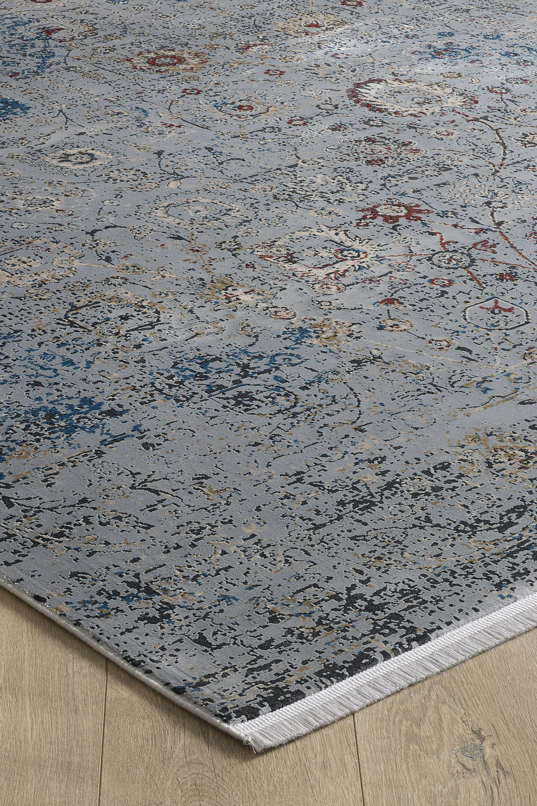 A close-up of the Miniature Elegance Oushak Rug - Grey - 2084F, a machine-made rug in blue with a distressed, abstract floral pattern and fringed edges, shown on a light wooden floor.