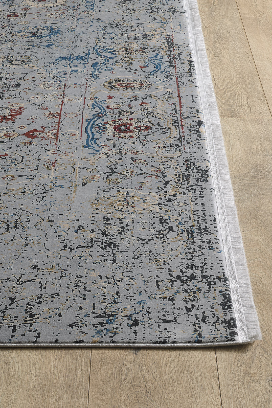 A close-up of the Miniature Elegance Oushak Rug - Grey - 2084F, a machine-made modal silk-polyester rug with distressed abstract blue, red, gold, and black patterns on a light background and fringed edges, shown on a light wood floor.