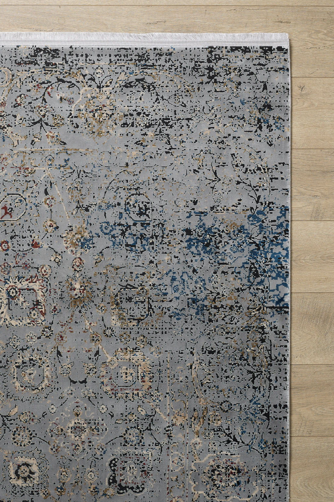 The upper right corner of the Miniature Elegance Oushak Rug - Grey - 2084F is shown on a light wooden floor, revealing its faded blue, beige, and gold floral and geometric design on a grey background.