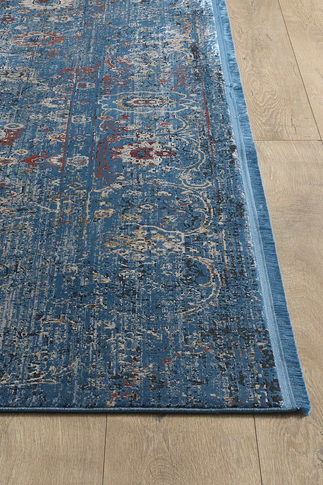 A close-up of the Miniature Elegance Oushak Rug - Navy - 2084E, a rectangular blue area rug with an intricate distressed pattern in blue, gray, and red hues, is shown on a light wood floor with its machine-made edge and texture visible.