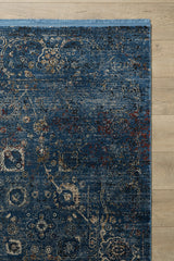 A close-up of the Miniature Elegance Oushak Rug - Navy - 2084E, a machine-made blue area rug featuring intricate beige and red floral and geometric patterns, with a blue fringe along the top edge, displayed on a light wooden floor.