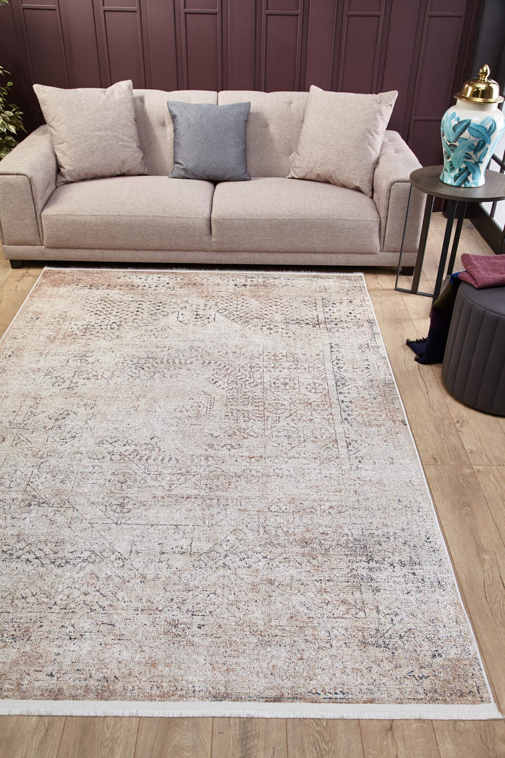 A beige sofa with three cushions sits against a dark mauve wall. On the wooden floor is the Misty Mosaic - Washable Rug - JR1690 (Custom Size), cream and beige in color, with a round side table and decorative jar on the right.