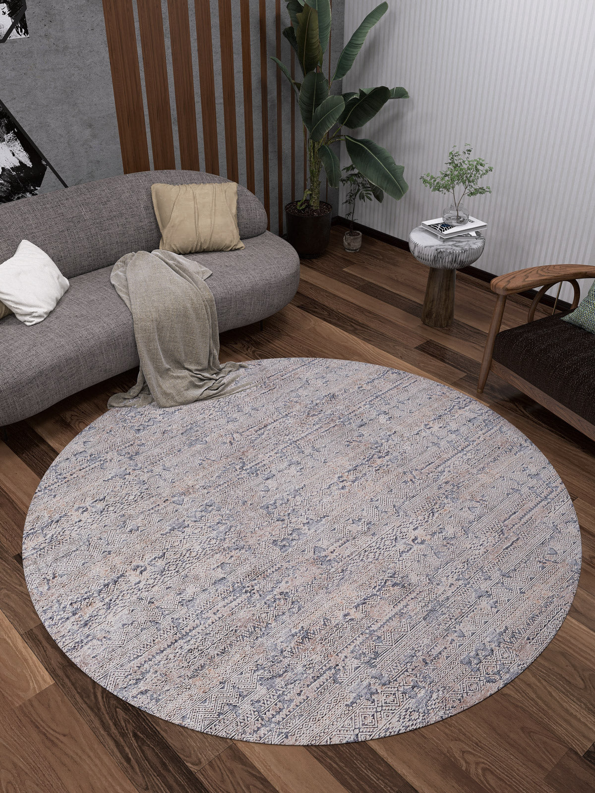 A modern living room showcases the Modern Enigma - Machine-Washable Rug JR1883 on wooden floors, paired with a curved gray sofa, soft cushions and throw, wooden chair, potted plant, and a small side table topped with greenery.