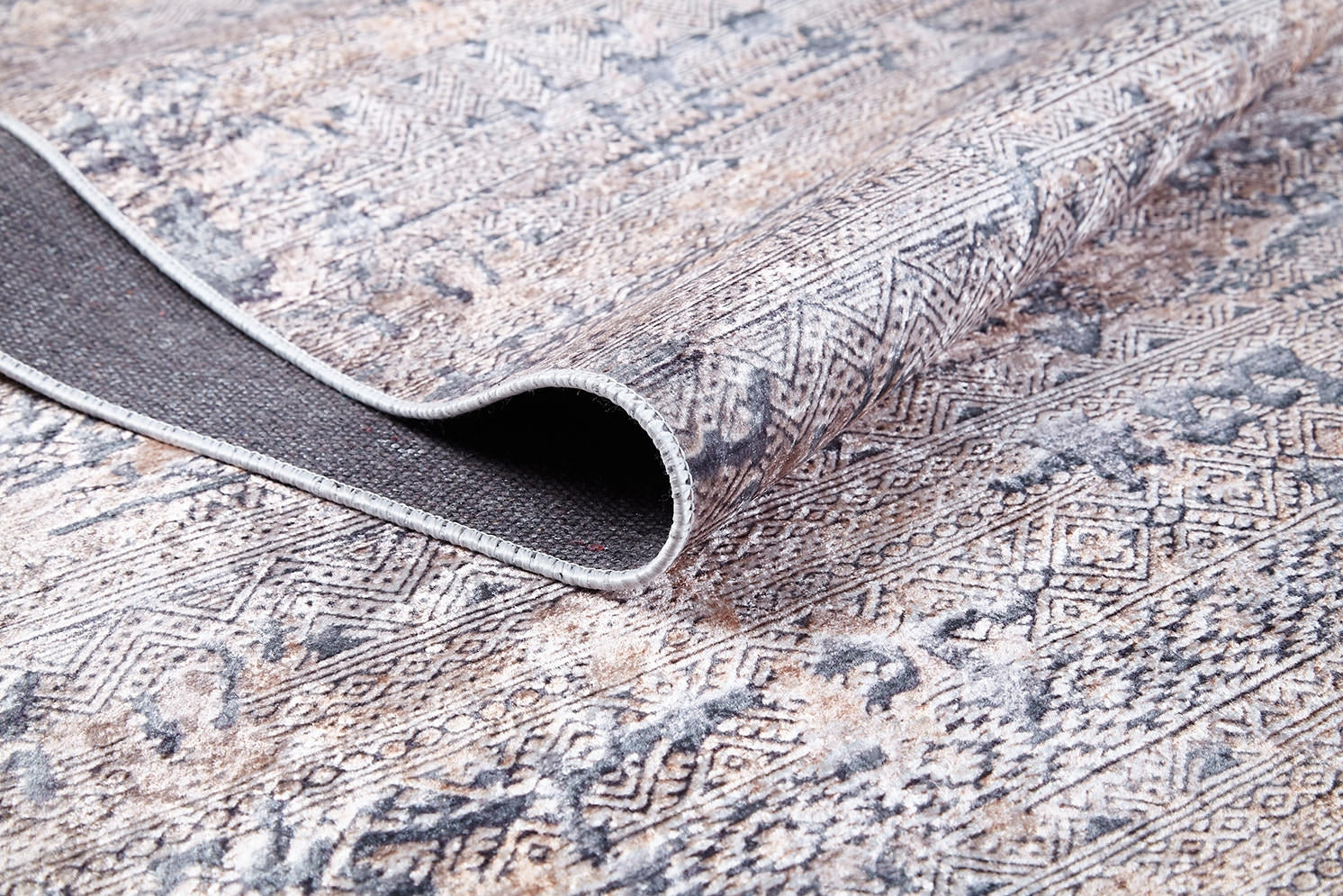 A close-up of the Modern Enigma - Machine-Washable Rug JR1883 (Custom Size) shows its geometric and ornate beige, gray, and brown patterns with a folded corner revealing the backing—ideal for high-traffic areas.