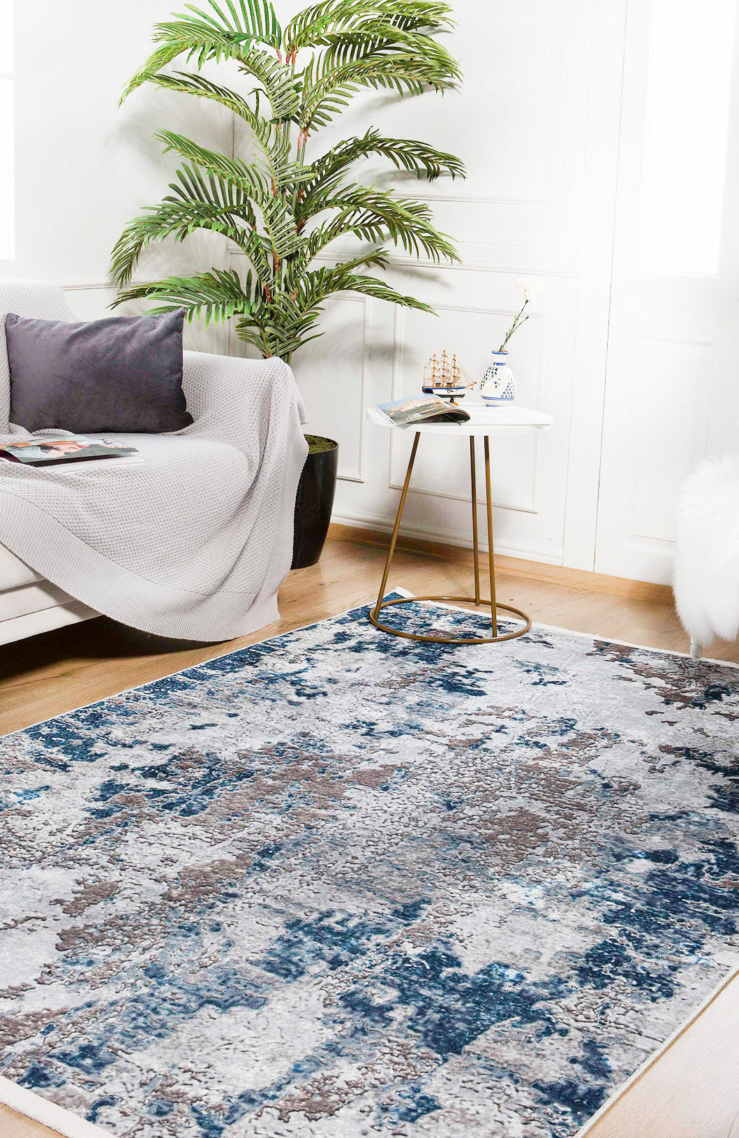 A modern living room featuring the Modern Melody - Washable Rug - JR586 (Custom Size) in blue and gray, a white sofa with gray pillows and blanket, a small round table with a teacup and model ship, a potted palm, and bright window light.