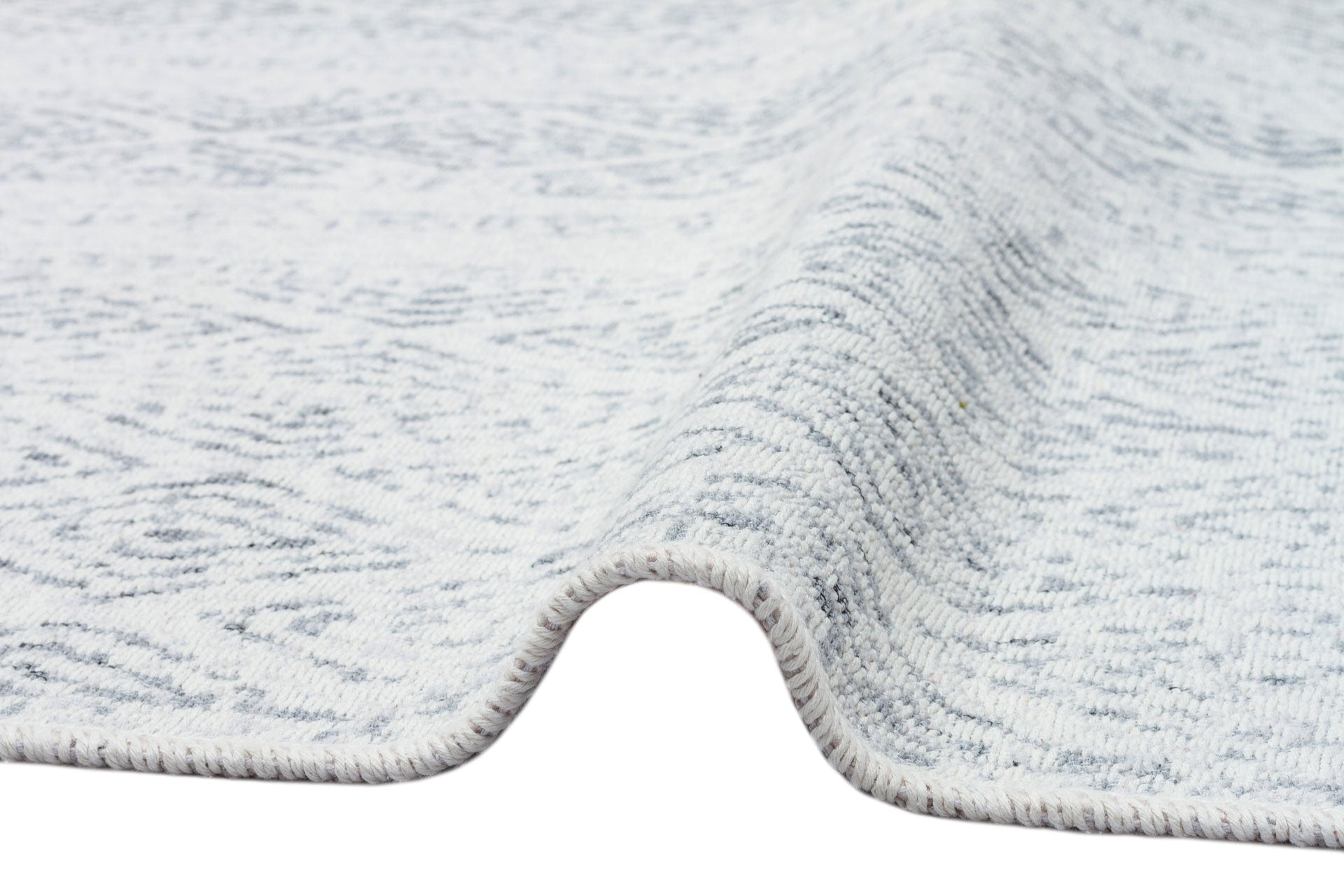 A close-up of the Modern Mist - Washable Double-Sided Kilims - AR01 rug, slightly lifted to reveal its texture, thickness, visible edge stitching, and a subtle pattern that enhances its modern style.