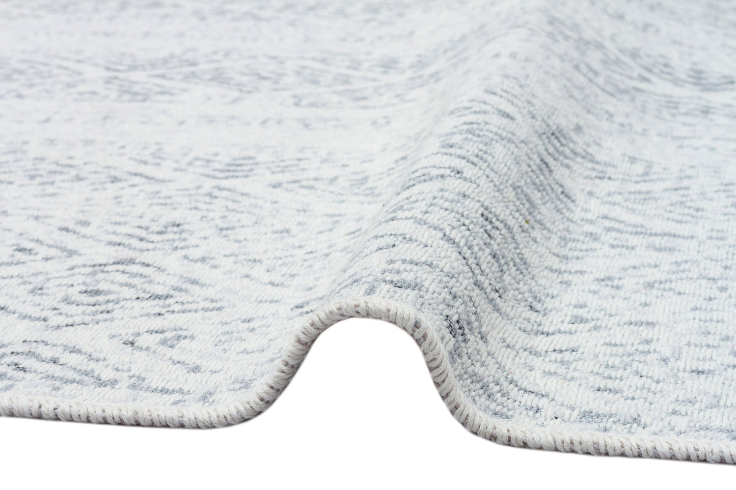A close-up of the Modern Mist - Washable Double-Sided Kilims - AR01 rug, slightly lifted to reveal its texture, thickness, visible edge stitching, and a subtle pattern that enhances its modern style.