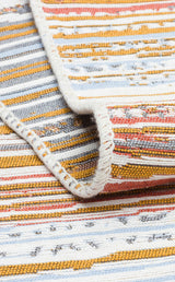 A close-up of the Mosaic Fade - Washable Double-Sided Kilims - AR35, featuring textured horizontal yellow, white, blue, and orange stripes with a folded corner displaying visible edge stitching.