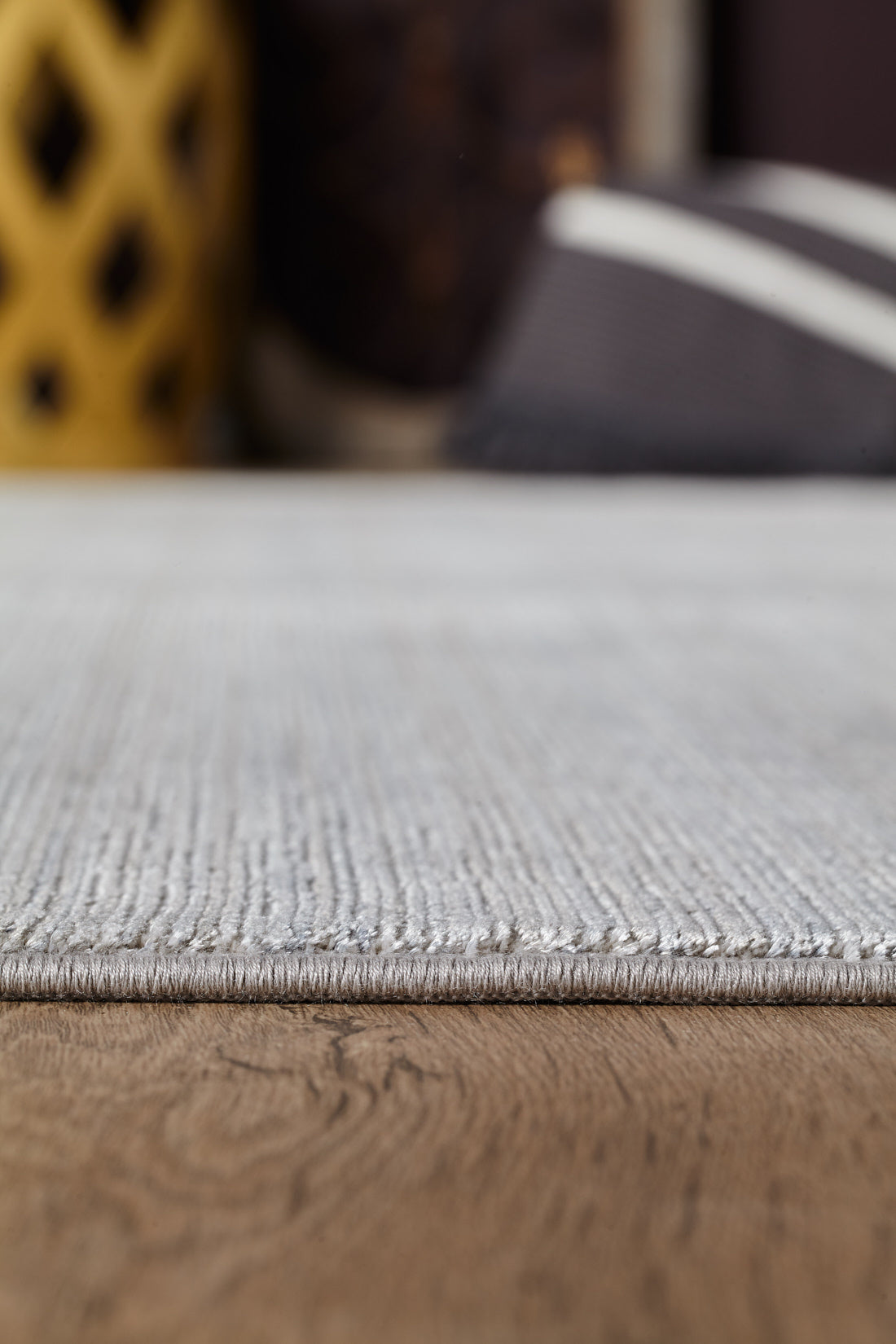 A close-up of the Muted Palette Grey Ombre Rug - EW2636 on a wooden floor, featuring its light gray texture, with blurred decorative objects and a yellow item in the background.