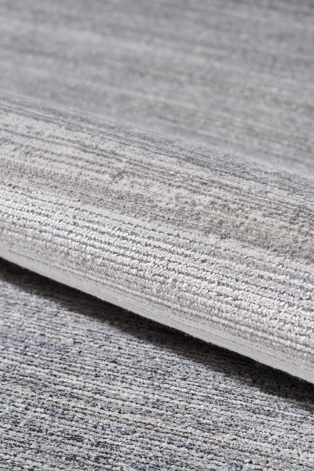 A close-up of the Muted Palette Grey Ombre Rug - EW2636, a machine-made piece in textured grey and beige with subtle linear patterns, shown rolled on a matching carpeted surface.