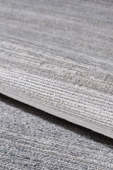 A close-up of the Muted Palette Grey Ombre Rug - EW2636, a machine-made piece in textured grey and beige with subtle linear patterns, shown rolled on a matching carpeted surface.