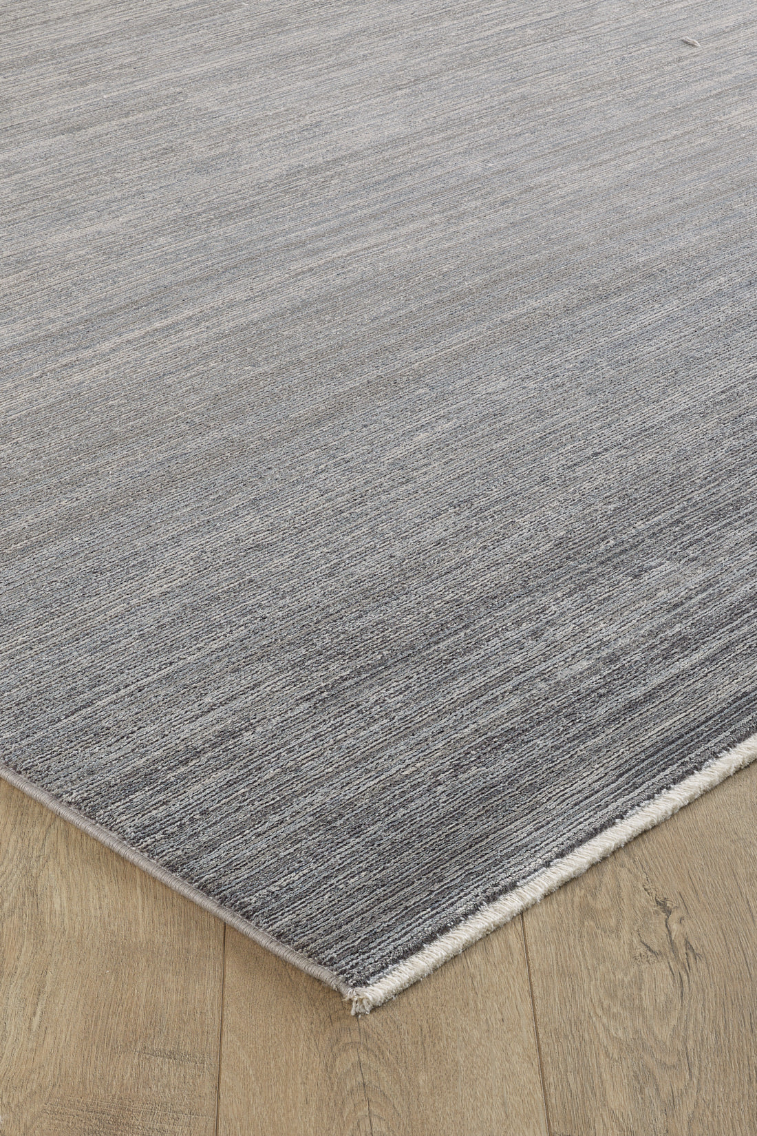 A close-up of the Muted Palette Grey Ombre Rug - EW2636, a modern machine-made piece with subtle linear patterns and a fringed edge, displayed on a light wooden floor in elegant neutral tones.
