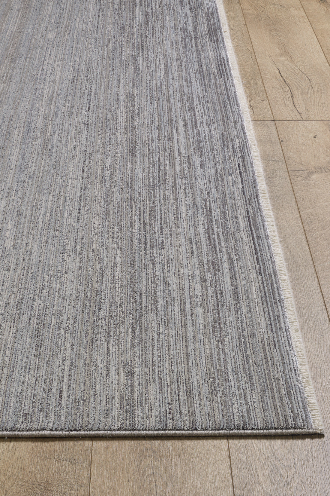 A close-up of the Muted Palette Grey Ombre Rug - EW2636, featuring gray and white vertical stripes, a subtle textured pattern, and fringed edges on the short sides, placed on a light wooden floor.