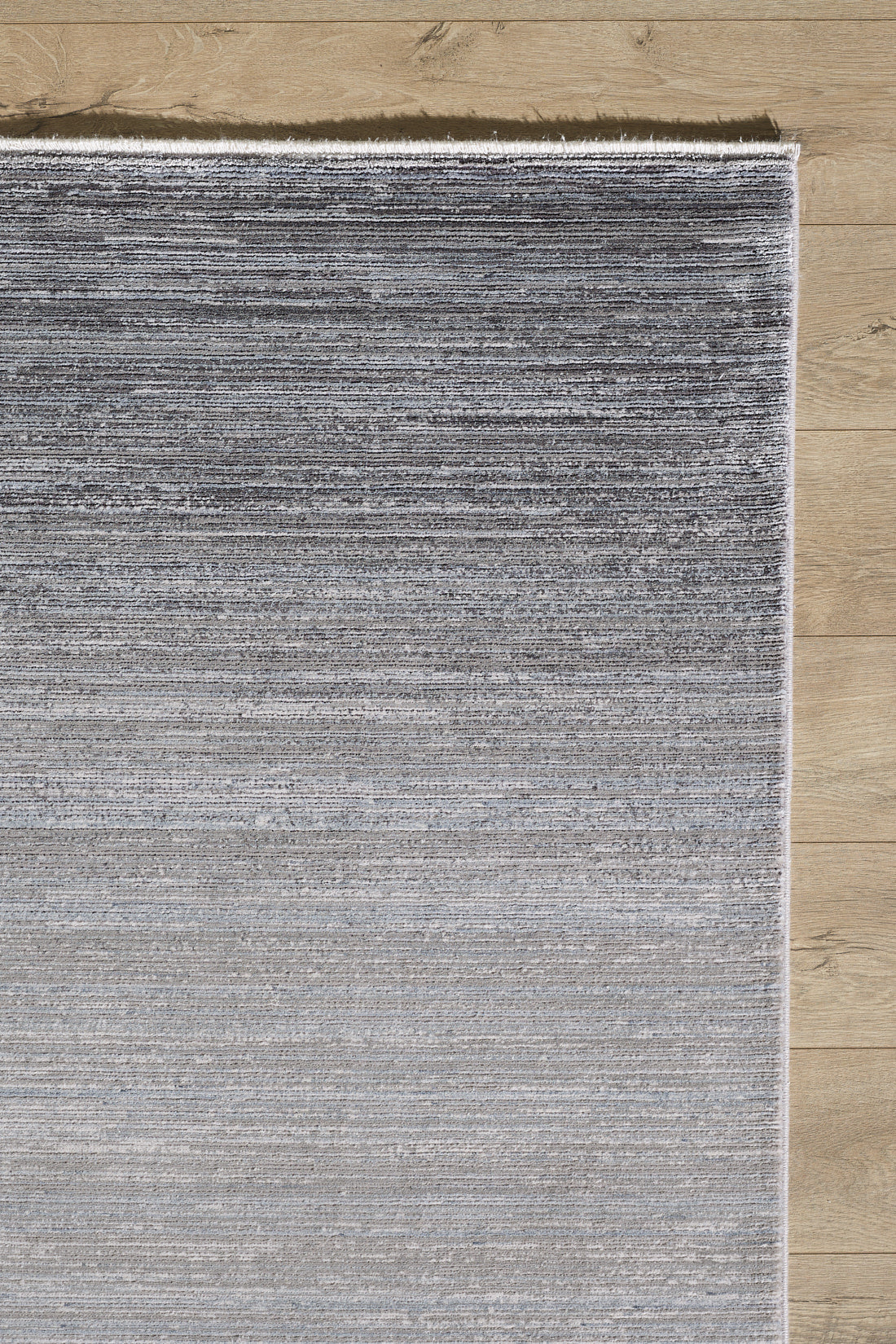 A close-up of the Muted Palette Grey Ombre Rug - EW2636 shows its textured, machine-made fibers forming a subtle striped gradient from dark to light gray, placed on a light wood floor.