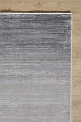 A close-up of the Muted Palette Grey Ombre Rug - EW2636 shows its textured, machine-made fibers forming a subtle striped gradient from dark to light gray, placed on a light wood floor.