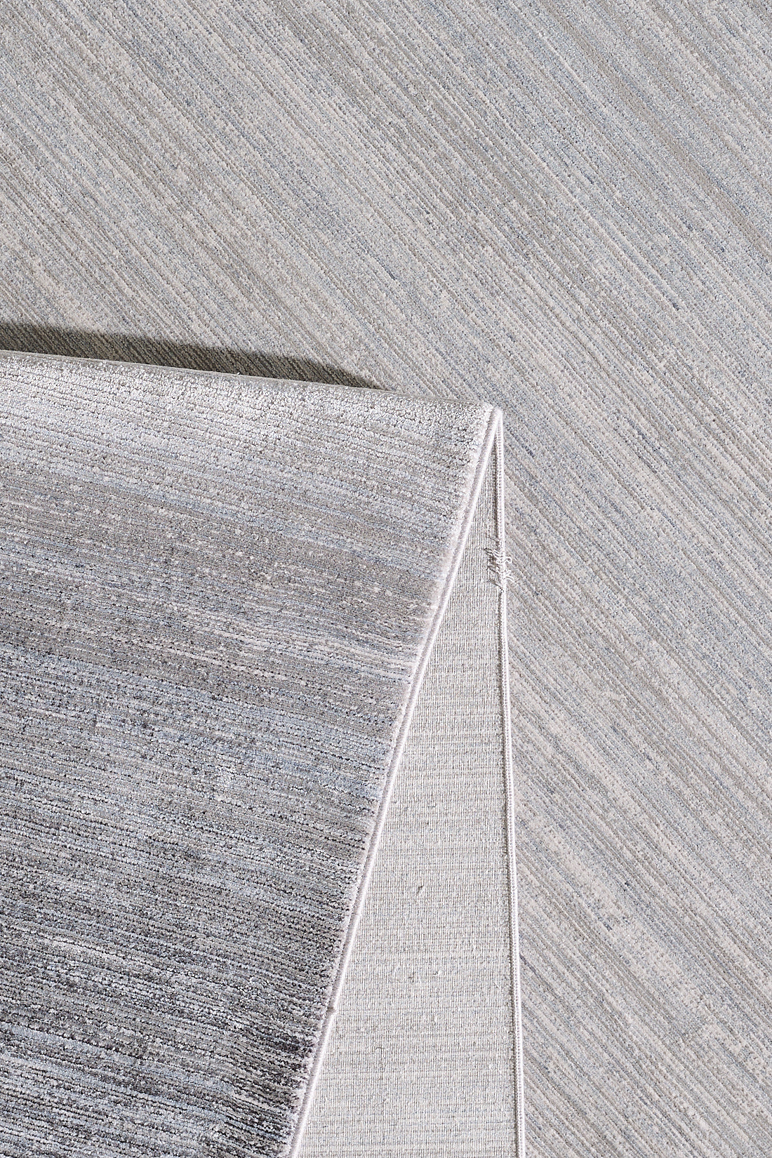 A close-up view of the Muted Palette Grey Ombre Rug - EW2636 shows its textured surface with a folded corner, highlighting the weave and subtle color variations in this modern, light grey neutral-toned rug.