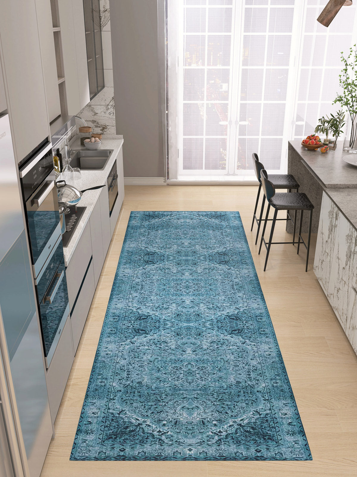 A modern kitchen with light cabinetry and appliances features the Mystic Mosaic - Washable Rug - JRN1019, a blue patterned runner on a light wood floor. A bar counter with black stools sits by tall windows that fill the space with natural light.