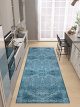 A modern kitchen with light cabinetry and appliances features the Mystic Mosaic - Washable Rug - JRN1019, a blue patterned runner on a light wood floor. A bar counter with black stools sits by tall windows that fill the space with natural light.