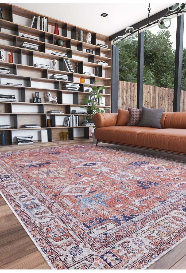 A modern living room features the Mystral Kilim-Design Coral Washable Rug - LCC3015, ideal for high-traffic areas, a brown leather sofa with cushions, a tall bookshelf with books and decor, and floor-to-ceiling windows overlooking greenery.