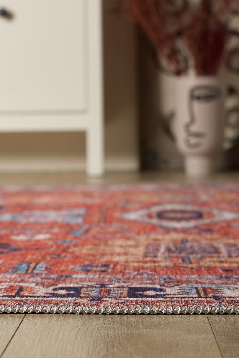 A close-up of the Mystral Kilim-Design Coral Washable Rug - LCC3015 with blue patterns on a wooden floor. A white cabinet and an abstract face vase with dried plants add style—ideal for high-traffic spaces.