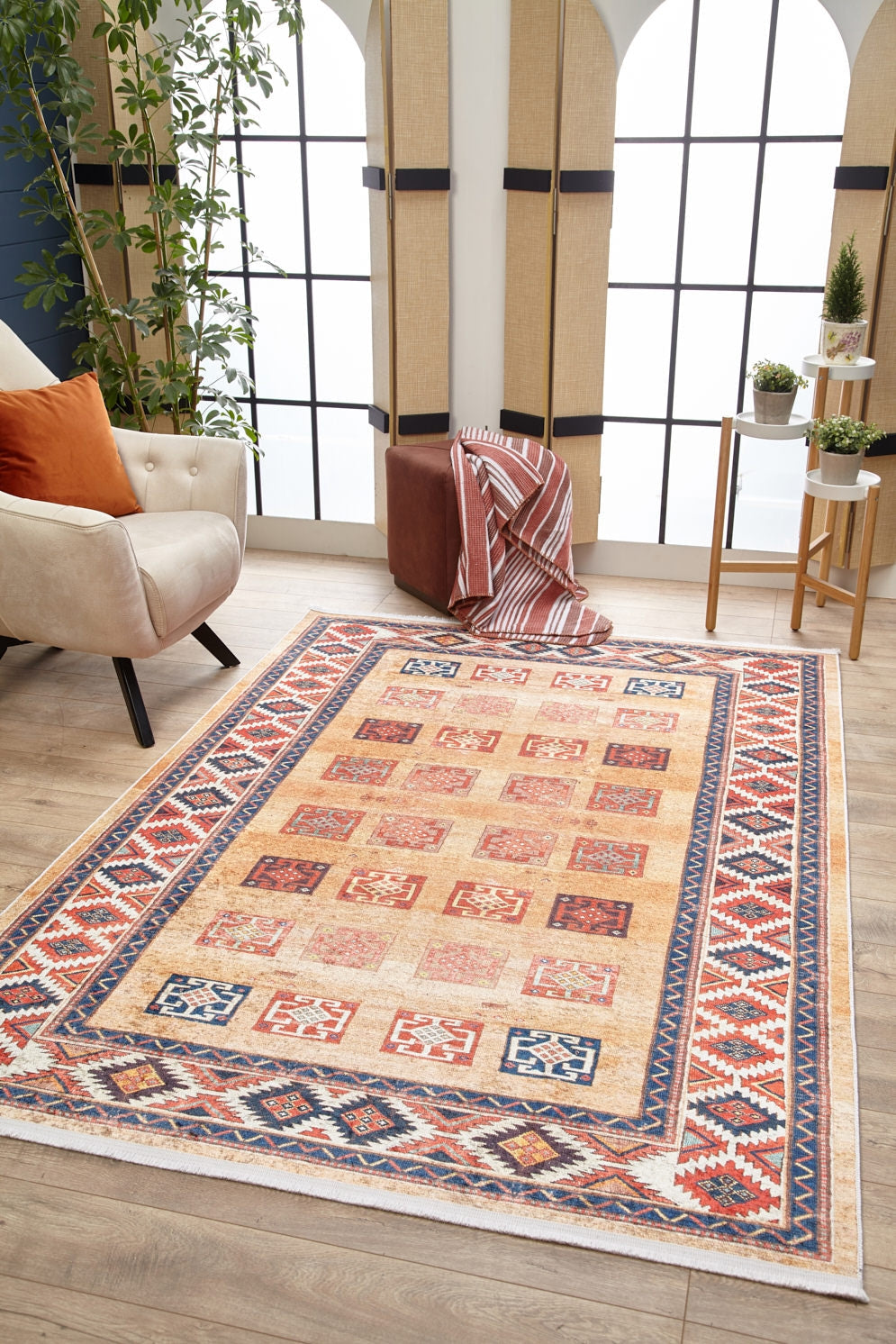 A living room features the Navajo Nights - Washable Rug - JR1758 (Custom Size), a cream armchair with orange cushions, a plant, brown ottoman draped with a striped blanket, and small side table with potted plants by large arched windows.