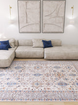A modern living room with a light beige sectional sofa, two blue accent pillows, the Nexa Bordered Beige Washable Rug (LCC3009), abstract wall art, and two wall-mounted lights. The decor is minimalist and neutral-toned.