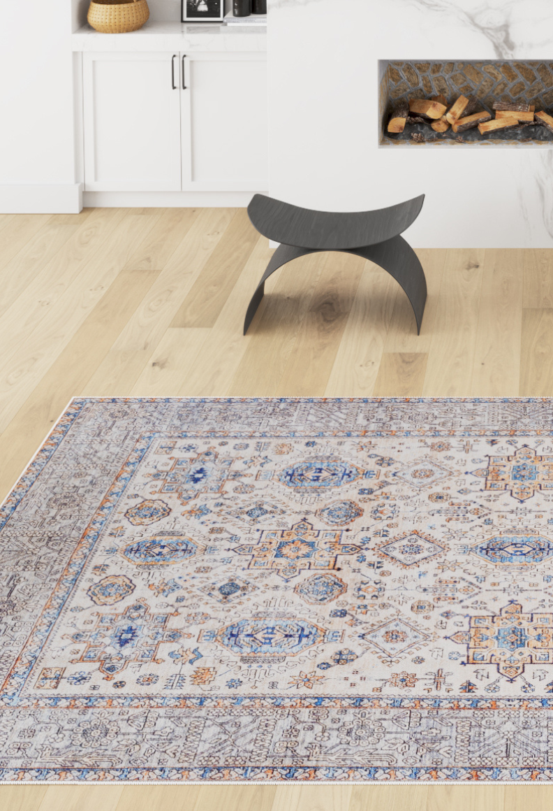 A modern living room with light wood floors, the Nexa Bordered Beige Washable Rug (LCC3009) featuring blue and orange accents, a black curved stool, fireplace with stacked logs, and white cabinets topped with a woven basket.