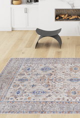 A modern living room with light wood floors, the Nexa Bordered Beige Washable Rug (LCC3009) featuring blue and orange accents, a black curved stool, fireplace with stacked logs, and white cabinets topped with a woven basket.