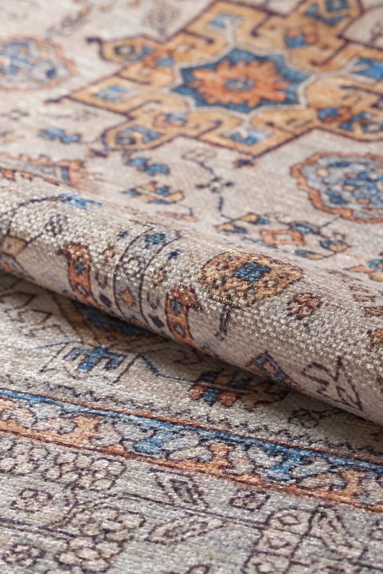 A close-up of the Nexa Bordered Beige Washable Rug - LCC3009 (Custom Size), showing intricate floral and geometric patterns in beige, blue, gold, and brown, with a folded edge highlighting its texture—ideal for high-traffic spaces.