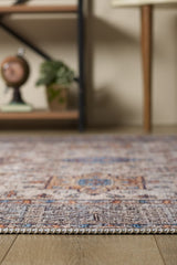 A close-up of the Nexa Bordered Beige Washable Rug - LCC3009 (Custom Size) on a wooden floor, ideal for high-traffic areas, with a shelf in the background holding a globe and potted plant, plus part of a table with wooden legs.