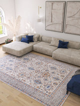 A spacious living room with a large beige sectional, blue accent pillows, the Nexa Bordered Beige Washable Rug - LCC3009 (Custom Size) for high-traffic areas, abstract wall art, a modern pendant light, and pampas grass in one corner.