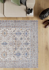 A beige couch with green pillows sits next to the Nexa Bordered Beige Washable Rug - LCC3009 (Custom Size), featuring blue, yellow, and gray designs—ideal for high-traffic areas—on a light wood floor with brown shoes, a black bag, and binoculars nearby.