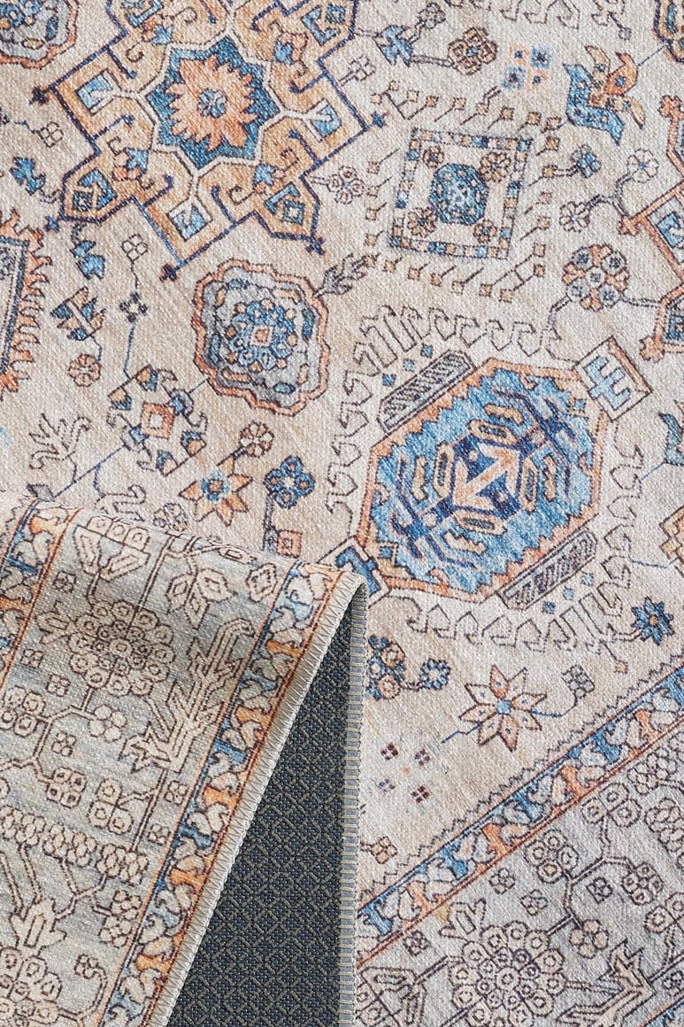 A close-up of the Nexa Bordered Beige Washable Rug - LCC3009 (Custom Size) shows intricate blue, orange, and beige geometric and floral patterns. A folded corner reveals its textured non-slip backing—ideal for high-traffic areas.