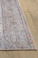 A close-up of the Nexa Bordered Beige Washable Rug - LCC3009 (Custom Size) shows its intricate floral and geometric patterns in blue, gray, and beige with orange-accented borders, laid on a light wood floor—perfect for high-traffic areas.