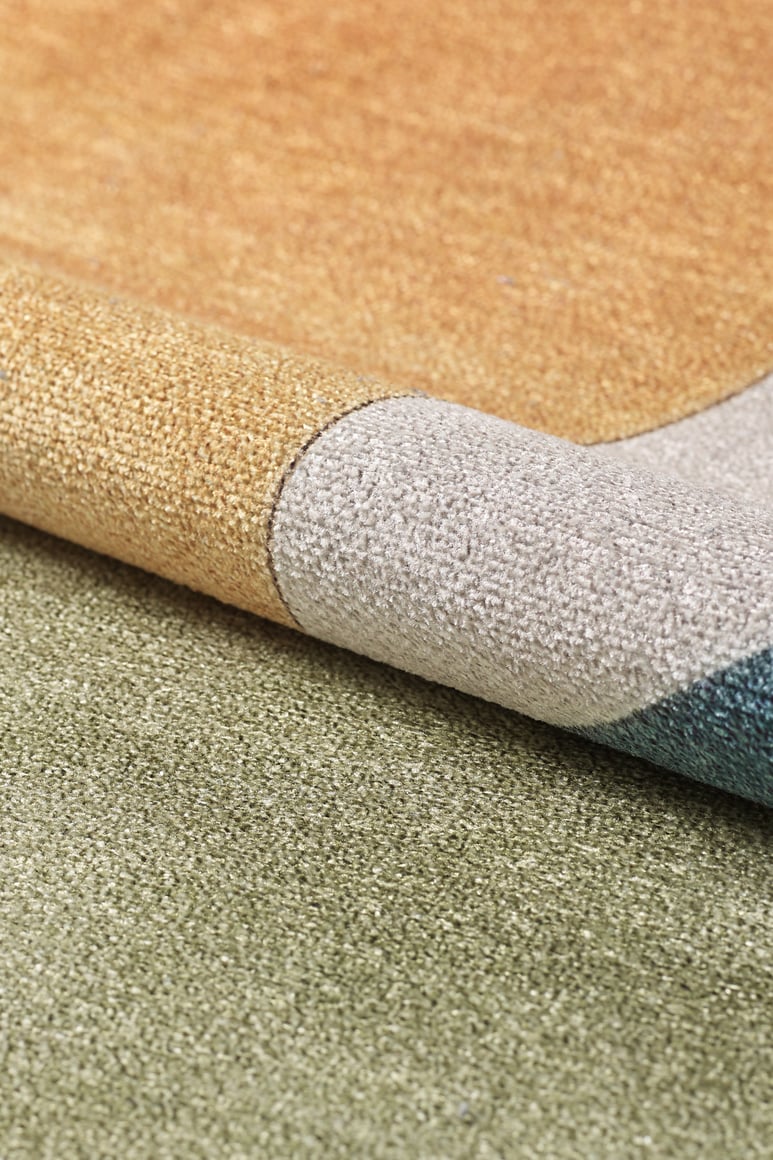 The Nexus Geometric Contemporary Washable Rug - MDJ006, in gold, gray, blue, and green tones, is shown slightly folded on a green carpet—an ideal choice for high-traffic spaces.