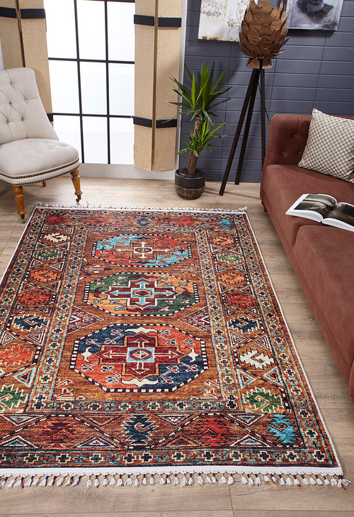 Bild 1 in Galerieansicht laden. A living room with the Nomadic Oasis - Washable Rug - JR5086 (Custom Size), featuring colorful geometric designs—ideal for high-traffic areas—plus a brown sofa with an open magazine, a white tufted chair, tall potted plant, and sheer-curtained windows.